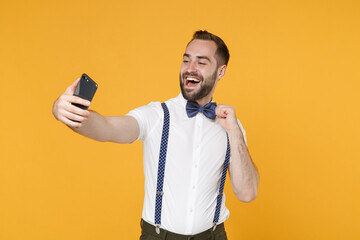 Funny cheerful laughing young bearded man 20s wearing white shirt bow-tie suspender posing standing doing selfie shot on mobile phone isolated on bright yellow color wall background studio portrait.