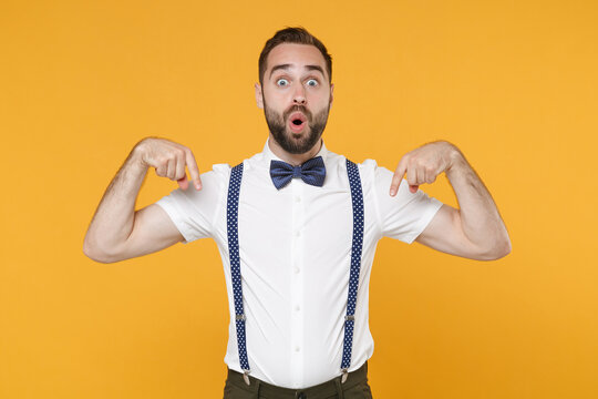 Shocked amazed young bearded man 20s in white shirt bow-tie suspender posing standing pointing index fingers down on mock up copy space isolated on bright yellow color wall background studio portrait.