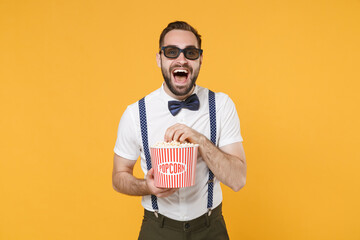 Laughing young bearded man 20s wearing white shirt bow-tie suspender 3d glasses posing watching movie film, holding bucket of popcorn isolated on bright yellow color wall background studio portrait.