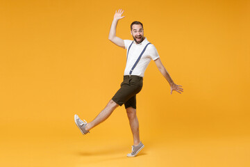 Fototapeta na wymiar Full length side view portrait of excited young bearded man 20s wearing white shirt suspender shorts posing rising leg spreading hands looking camera isolated on bright yellow color background studio.