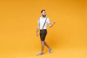 Fototapeta na wymiar Full length side view portrait excited young bearded man 20s wearing white shirt suspender shorts posing pointing index finger aside on mock up copy space isolated on yellow color background studio.