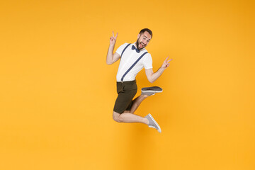 Full length side view portrait of excited young bearded man 20s wearing white shirt suspender shorts posing jumping showing victory sign looking camera isolated on yellow color wall background studio.
