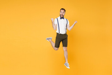 Fototapeta na wymiar Full length portrait of excited happy young bearded man 20s wearing white shirt suspender shorts posing jumping doing winner gesture looking camera isolated on bright yellow color background studio.