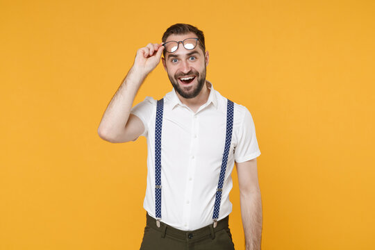 Excited surprised young bearded man 20s wearing white shirt suspender eyeglasses posing keeping mouth open looking camera isolated on bright yellow color wall background, studio portrait.