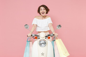 Excited surprised young brunette woman 20s wearing white summer clothes hold package bag with purchases after shopping sitting driving moped isolated on pastel pink colour background, studio portrait.