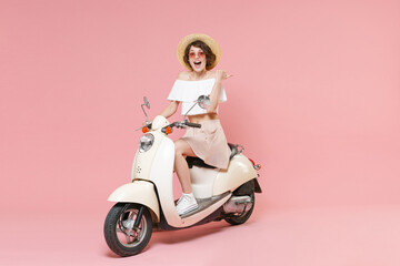 Fototapeta na wymiar Full length portrait of excited cheerful young woman in white summer clothes hat glasses point thumb aside on mock up copy space sitting driving moped isolated on pastel pink colour background studio.