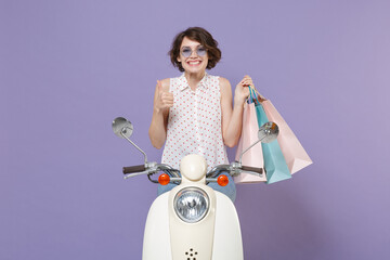 Smiling young brunette woman in white dotted shirt glasses hold package bag with purchases after shopping showing thumb up driving moped isolated on pastel violet colour background studio portrait.