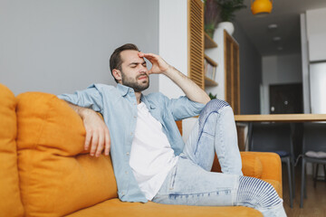Fototapeta premium Exhausted tired young bearded man wearing casual white t-shirt blue shirt keeping eyes closed put hand on head having headache sitting on couch resting relaxing spending time in living room at home.