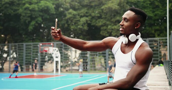 Handsome young African American guy sitting on basketball playground and making selfie photo on smartphone outdoor. Portrait of man athlete taking pictures on cellphone. Boys playing on background