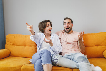 Shocked excited young couple two friends man woman 20s in casual clothes sitting on couch hugging spreading hands pointing index finger aside resting relaxing spending time in living room at home.