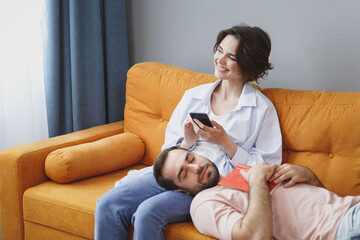 Funny smiling young couple two friends man woman 20s wearing casual clothes sitting lying on couch using mobile cell phone reading book sleeping resting relaxing spending time in living room at home.