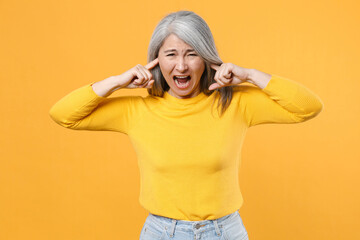 Crazy frustrated displeased gray-haired asian woman wearing casual clothes standing covering ears with fingers screaming looking camera isolated on bright yellow colour background, studio portrait.