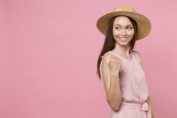 Side view of smiling charming young brunette woman 20s wearing pink summer dotted dress hat posing pointing thumb aside on mock up copy space isolated on pastel pink color background studio portrait.