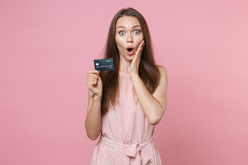 Shocked young brunette woman 20s wearing pink summer dotted dress posing holding in hand credit bank card put hand on cheek looking camera isolated on pastel pink color background studio portrait.