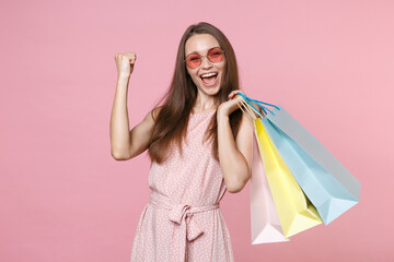 Happy young brunette woman 20s wearing pink summer dotted dress eyeglasses hold package bag with purchases after shopping doing winner gesture isolated on pastel pink color background studio portrait.