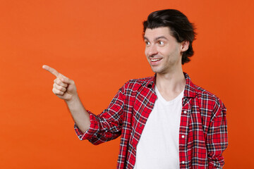 Funny smiling young brunet man 20s wearing white t-shirt red checkered shirt posing standing pointing index finger aside on mock up copy space isolated on orange color wall background studio portrait.