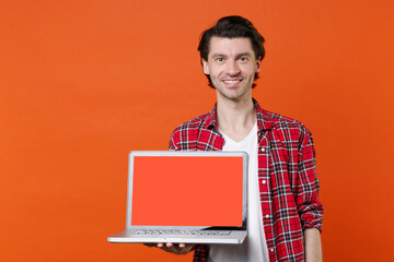 Smiling young man 20s in white t-shirt red checkered shirt posing hold laptop pc computer with blank empty screen mock up copy space looking camera isolated on orange wall background studio portrait.