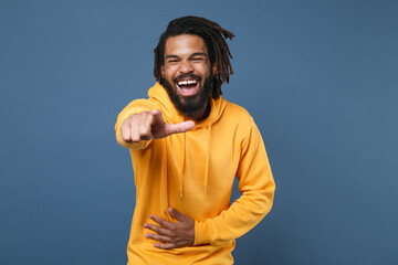 Naklejka premium Laughing young african american man guy in yellow streetwear hoodie posing isolated on blue background studio portrait. People lifestyle concept. Pointing index finger on camera, put hand on stomach.