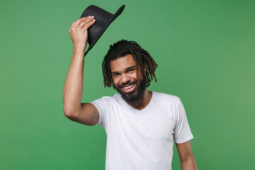 Fototapeta premium Funny smiling attractive young african american man guy with dreadlocks 20s in white casual t-shirt posing throwing black hat looking camera isolated on green color wall background studio portrait.