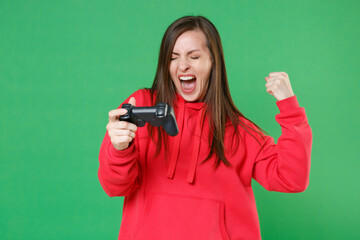 Screaming overjoyed young brunette woman 20s wearing bright red casual streetwear hoodie posing playing game with joystick doing winner gesture isolated on green color wall background studio portrait.