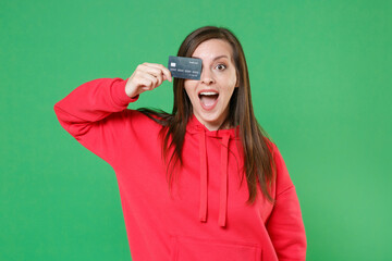 Surprised cheerful young brunette woman 20s wearing bright red casual streetwear hoodie posing covering eye with credit bank card keeping mouth open isolated on green color background studio portrait.