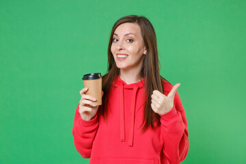 Smiling beautiful cheerful young brunette woman 20s wearing red casual streetwear hoodie posing showing thumb up hold paper cup of coffee or tea isolated on green color background studio portrait.