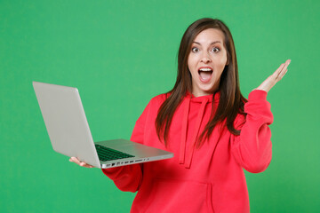 Surprised young brunette woman 20s in bright red casual streetwear hoodie posing working on laptop pc computer spreading hands keeping mouth open isolated on green color background studio portrait.