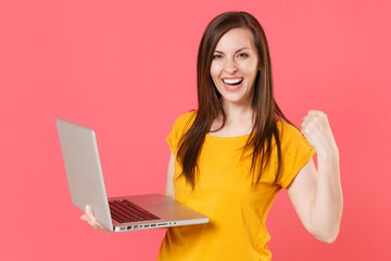 Naklejka premium Happy joyful young brunette woman 20s wearing yellow casual t-shirt posing and working on laptop pc computer doing winner gesture looking camera isolated on pink color wall background studio portrait.
