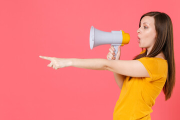 Side view of shocked young brunette woman in yellow casual t-shirt posing screaming in megaphone pointing index finger aside on mock up copy space isolated on pink color background studio portrait.