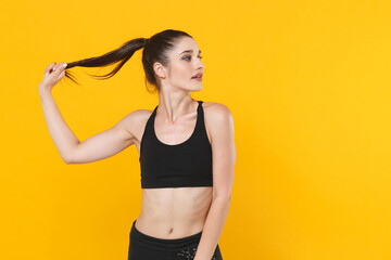 Fototapeta na wymiar Beautiful attractive young fitness sporty woman 20s wearing black sportswear posing working out training holding hair ponytail looking aside isolated on bright yellow color background studio portrait.