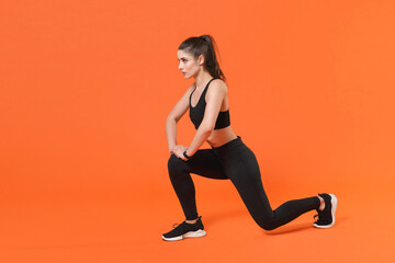 Fototapeta na wymiar Full length side view portrait of beautiful young fitness sporty woman in black sportswear posing training working out doing exercise lunge looking aside isolated on orange color background studio.