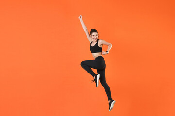 Full length side view of portrait of smiling young fitness sporty woman wearing black sportswear training working out jumping clenching fists rising hands isolated on orange color background studio.
