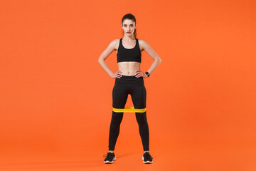 Fototapeta na wymiar Full length portrait of young fitness sporty woman in black sportswear training working out doing exercises with fitness gums standing with arms akimbo on waist isolated on orange background studio.