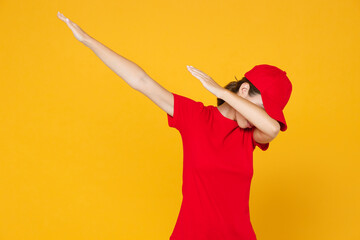 Delivery employee woman in red cap blank t-shirt uniform work courier in service during quarantine coronavirus covid-19 virus doing dab dance gesture hiding covering face isolated on yellow background