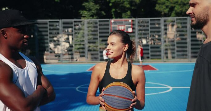 Close up portrait of mixed-races male and female basketball players standing on outdoor court and talking. Beautiful Caucasian girl and guy speaking with handsome African American man about game