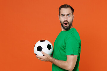 Side view worried young man football fan in green t-shirt cheer up support favorite team with soccer ball looking camera isolated on orange background studio. People sport leisure lifestyle concept.