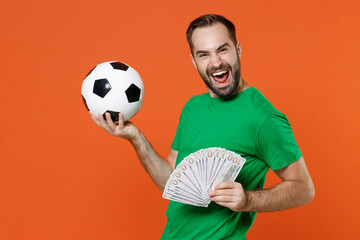 Happy young man football fan in green t-shirt cheer up support favorite team with soccer ball hold fan of cash money in dollar banknotes isolated on orange background. People sport leisure concept.