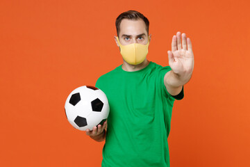 Young man football fan in green t-shirt face mask to safe from coronavirus support favorite team hold ball showing stop gesture with palm isolated on orange background. People sport leisure concept. - Powered by Adobe