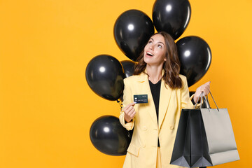 Funny young woman 20s in suit jacket hold package bags with purchases after shopping bunch of air balloons credit bank card looking up isolated on yellow background studio portrait. Black friday sale.