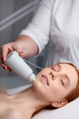 Naklejka premium redhead woman get face lifting by ultrasound using a special cosmetology device, lie on bed in beauty salon