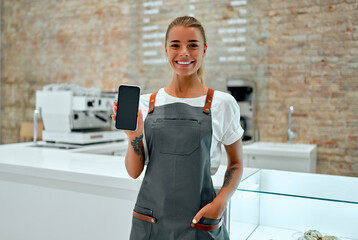 Young attractive woman barista stands at the counter in a coffee shop and smiles while showing a blank smartphone screen. - Powered by Adobe