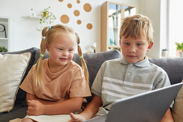 Portrait of cute girl with down syndrome smiling while looking at laptop screen and sitting on sofa...