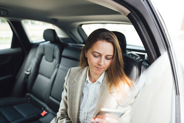 Young smarty business woman hold smartphone in hand while sits in car on backsit. Business lady using smartphone while traveling by car