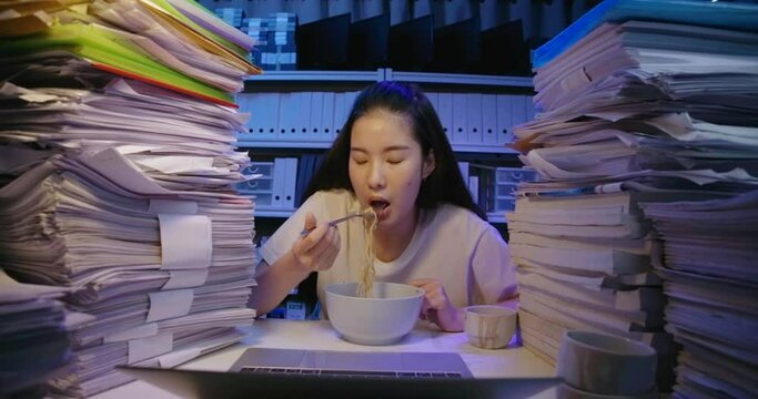 Hungry Young Asian Student Woman Is Sitting At Desk Cover With Stack Of Paperwork And Eating Instant Noodles For A Late Meal. Alone Tired Teen Girl Use Laptop While Studying Hard At Late Night.