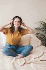 Young beautiful girl student in an orange T-shirt and blue jeans sits on bed with pillows and listens to an audiobook, music, podcast on headphones. Self-education concept at home during quarantine.