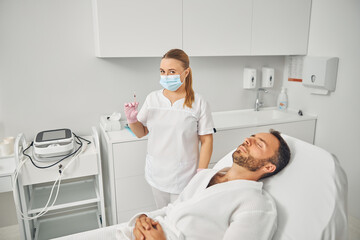 Female beautician standing beside young man in cosmetology cabinet