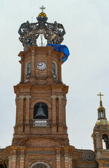 Puerto Vallarta, Mexico - April 25, 2008: Church of Our Lady of Guadalupe . Closeup of top of brown-red brick stone clock tower against light blue sky. Blue tarp and yellow globe add colors.