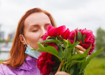 Revival after coronavirus. Redhead girl sniffs pink flowers with pulled down medicine mask. Recovery after coronavirus and flu. Sense of smell return. Discharge from hospital.