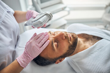 Handsome young man getting laser facial treatment in cosmetological clinic