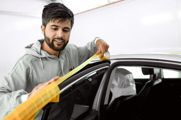 young caucasian car mechanic applying masking tape to a sanded down car before a paint job, in auto workshop. alonem wearing uniform